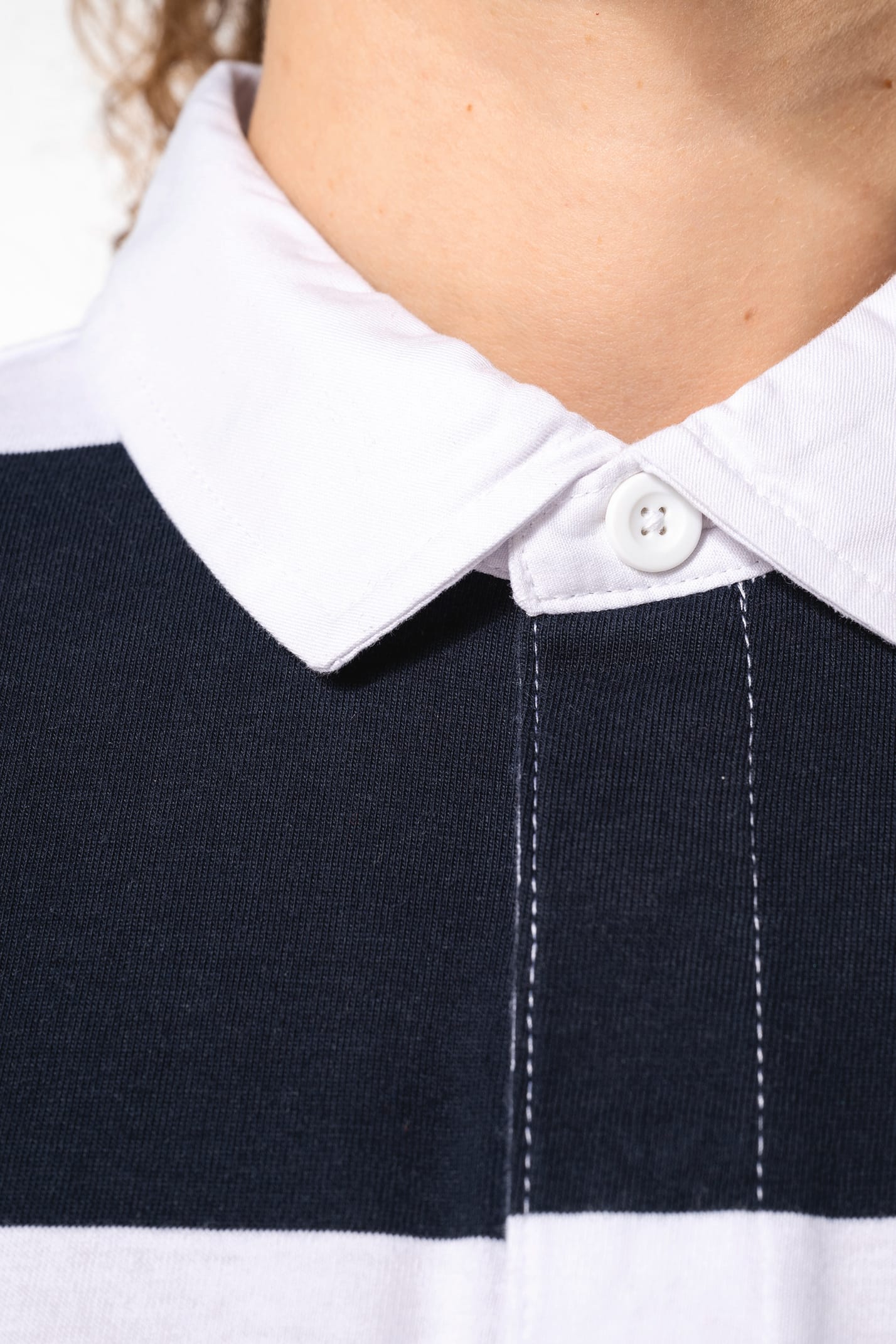 Jeune femme bouclée cheveux attachés qui porte un polo de rugby rayé blanc et bleu marine.