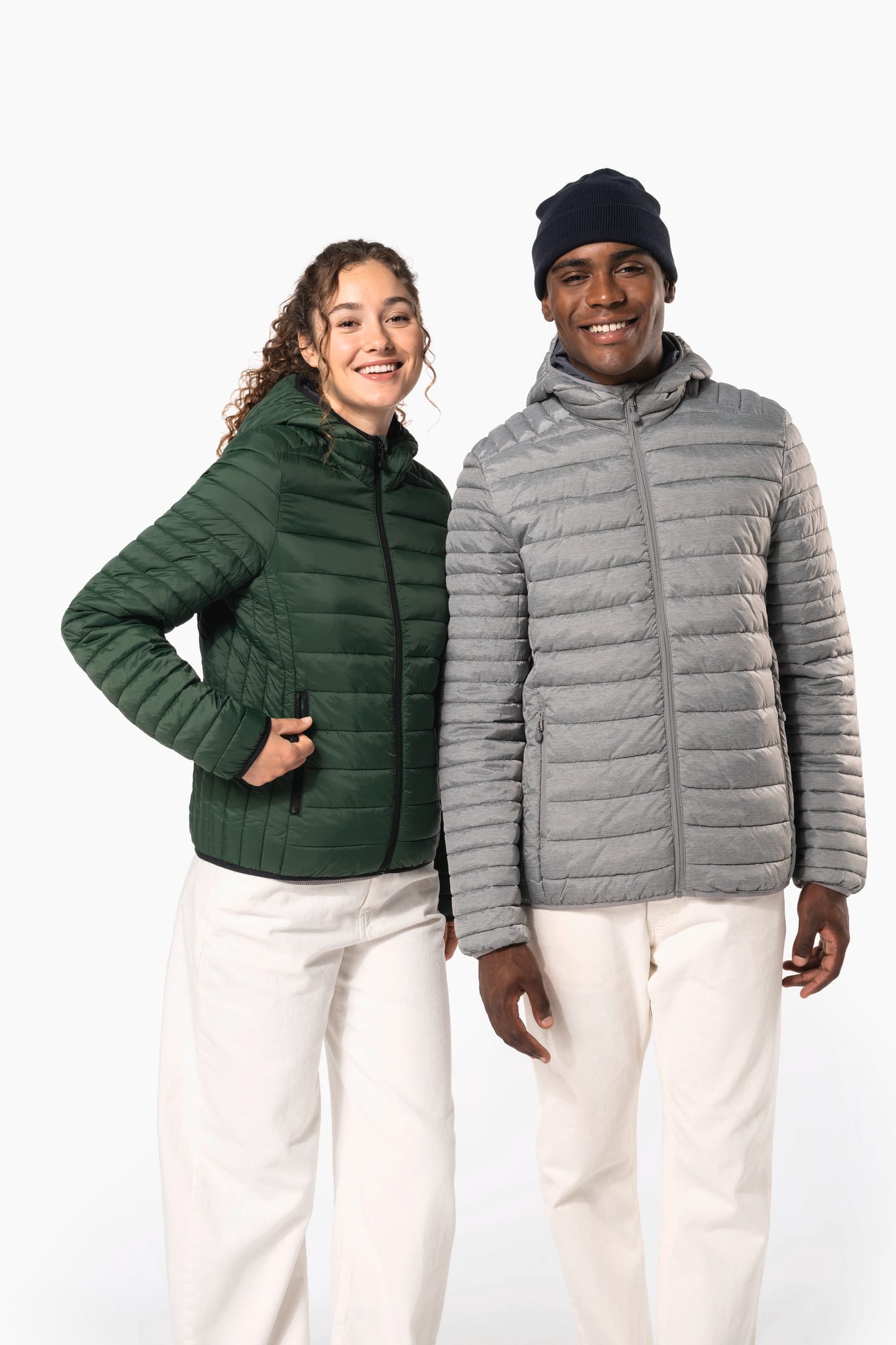Femme aux cheveux frisls attachés et homme brun avec bonnet noir qui portent des doudounes légères à capuches vert forêt et argenté.