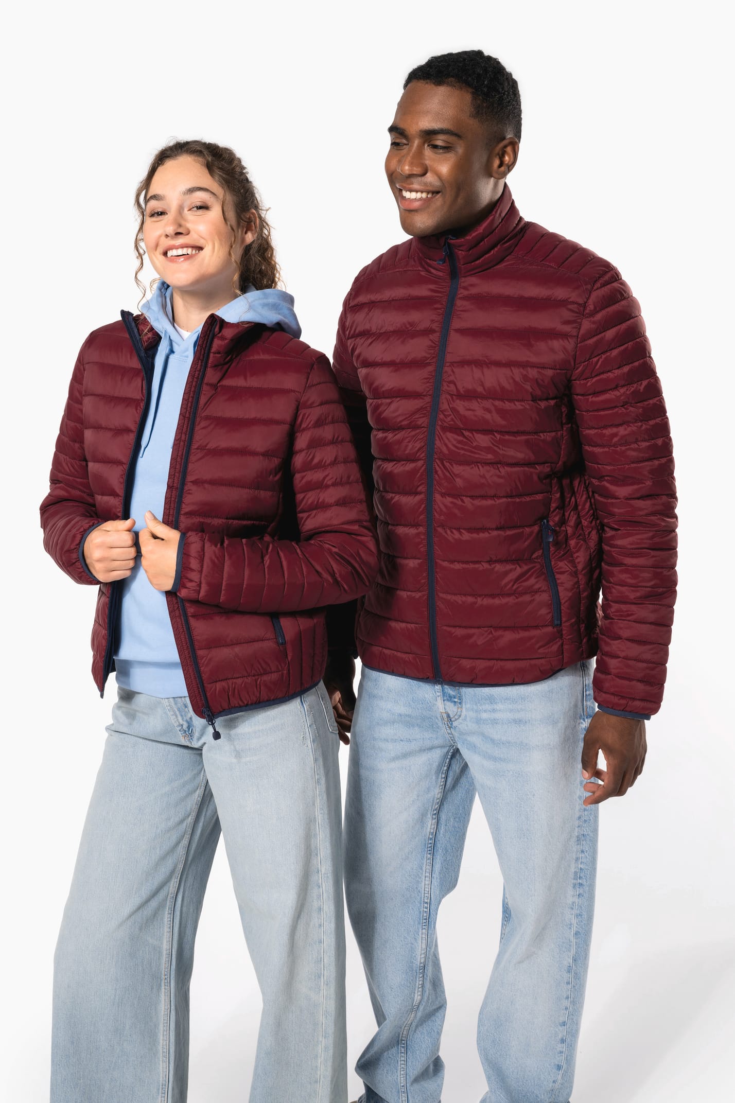 Jeune homme brun aux cheveux courts et jeune femme aux cheveux longs frisés attachés et sweat bleu clair qui portent des doudounes légères bordeaux.
