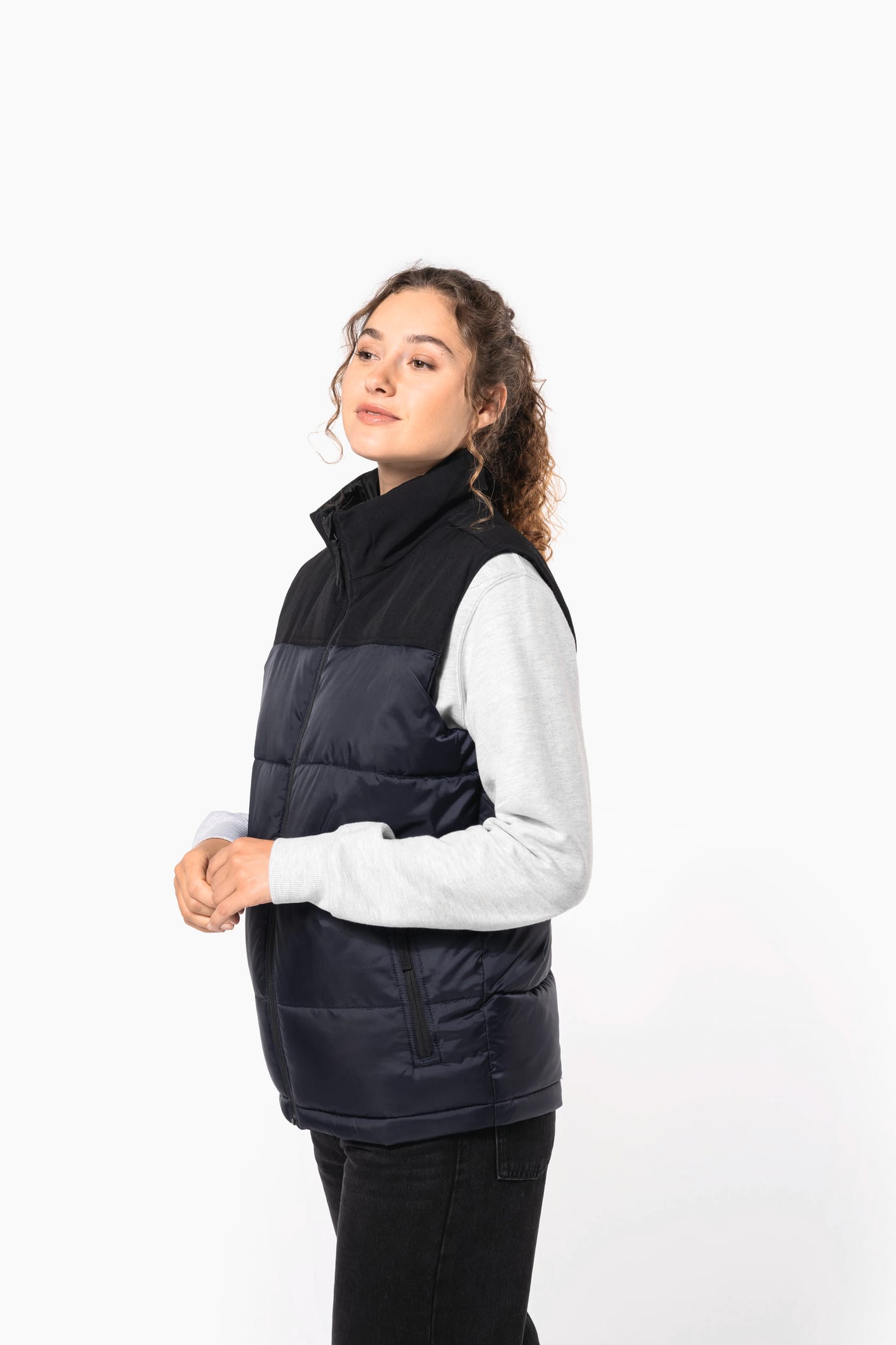Jeune femme châtain frisée avec queue de cheval, qui porte un bodywarmer matelassé bicolore marine et noir.