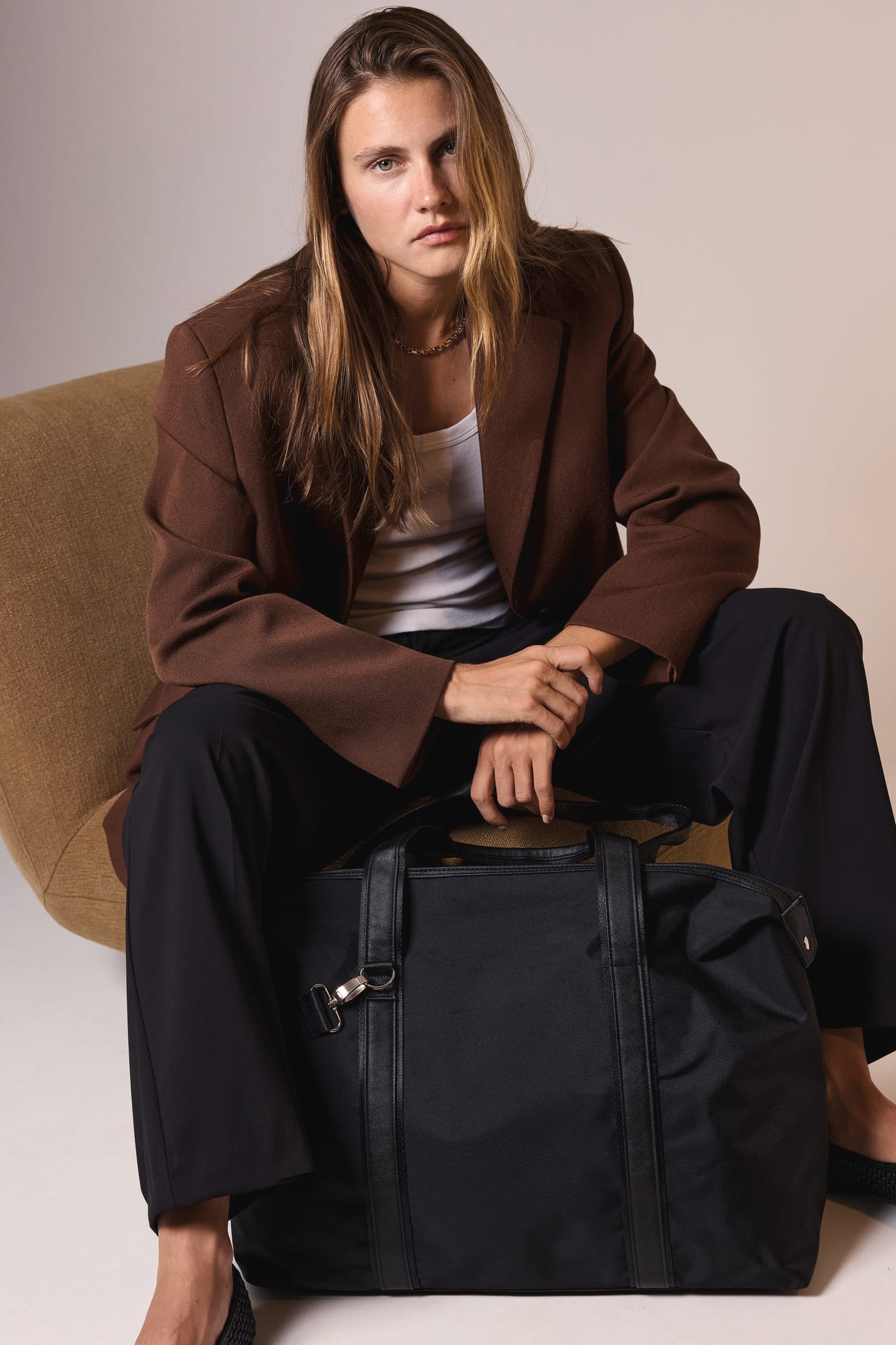 Jeune femme blonde aux cheveux longs avec un sac de voyage noir assise sur un fauteuil camel.