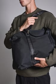Jeune homme debout qui tient fermement  un sac de voyage noir.