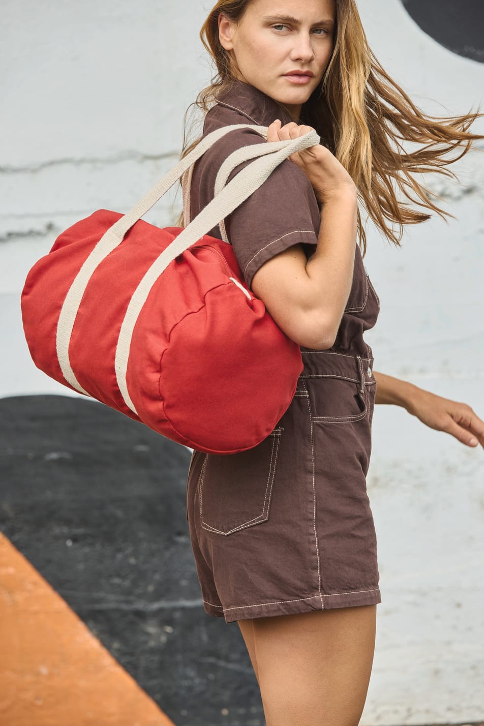 Belle jeune femme blonde aux cheveux long en salopette marron qui se promène avec un sac fourre-tout rouge.