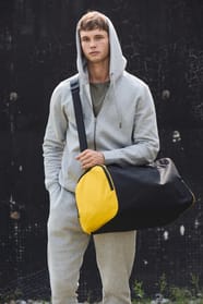 Jeune homme en jogging gris à capuche qui marche avec un sac de sport bicolore noir et jaune?