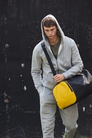 Jeune homme en jogging gris à capuche qui marche avec un sac de sport bicolore noir et jaune?
