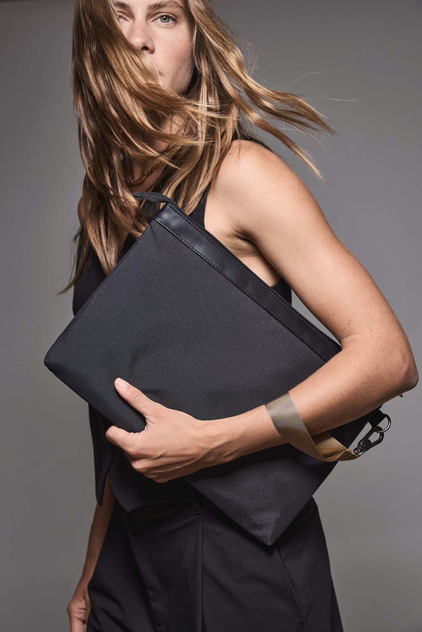 Jeune femme blonde aux cheveux longs qui porte un robe noire et qui tient une pochette pour documents et ordinateur portable noire.