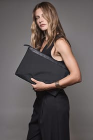 Jeune femme blonde aux cheveux longs qui porte un robe noire et qui tient une pochette pour documents et ordinateur portable noire.