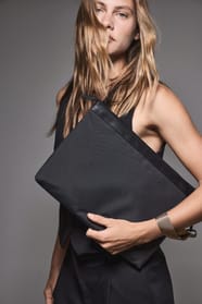 Jeune femme blonde aux cheveux longs qui porte un robe noire et qui tient une pochette pour documents et ordinateur portable noire.