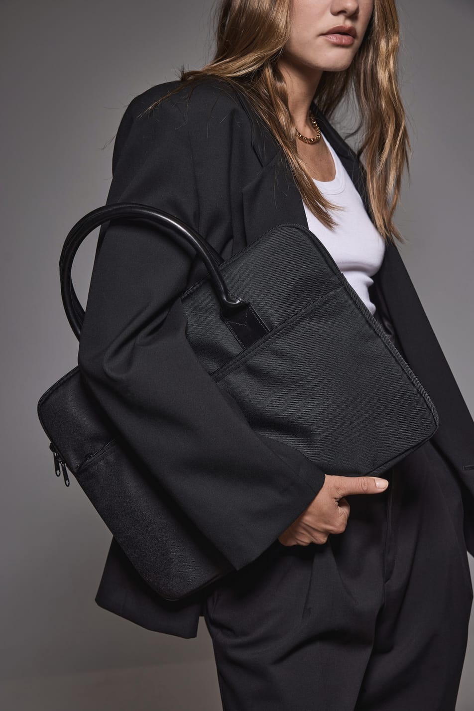 Jeune femme blonde aux cheveux longs en tenue business noire et blanche et tenant un sac porte-ordianteur noir.