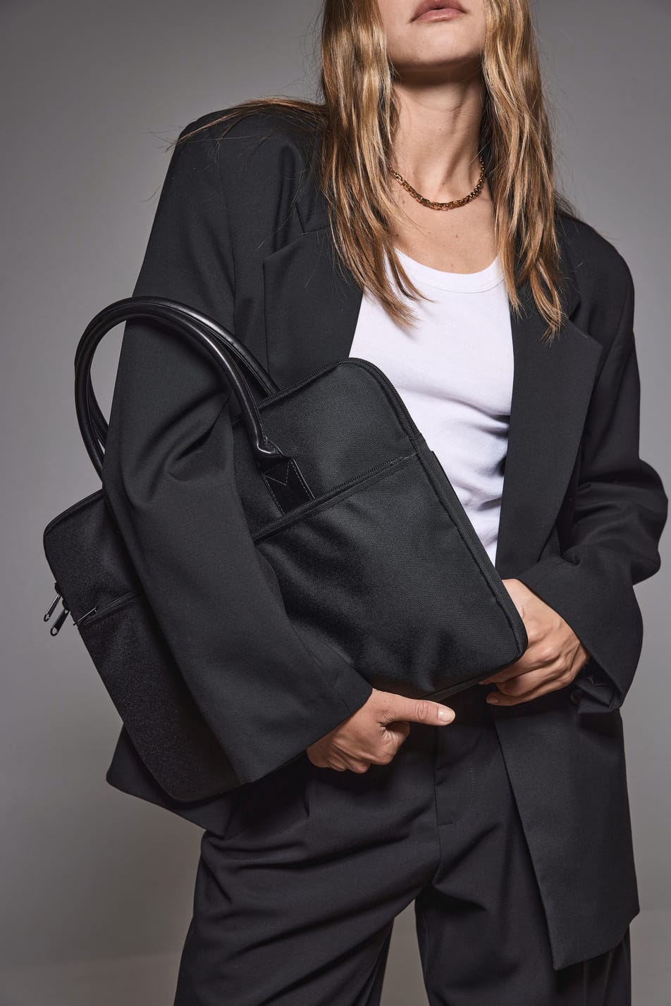Jeune femme blonde aux cheveux longs en tenue business noire et blanche et tenant un sac porte-ordianteur noir.