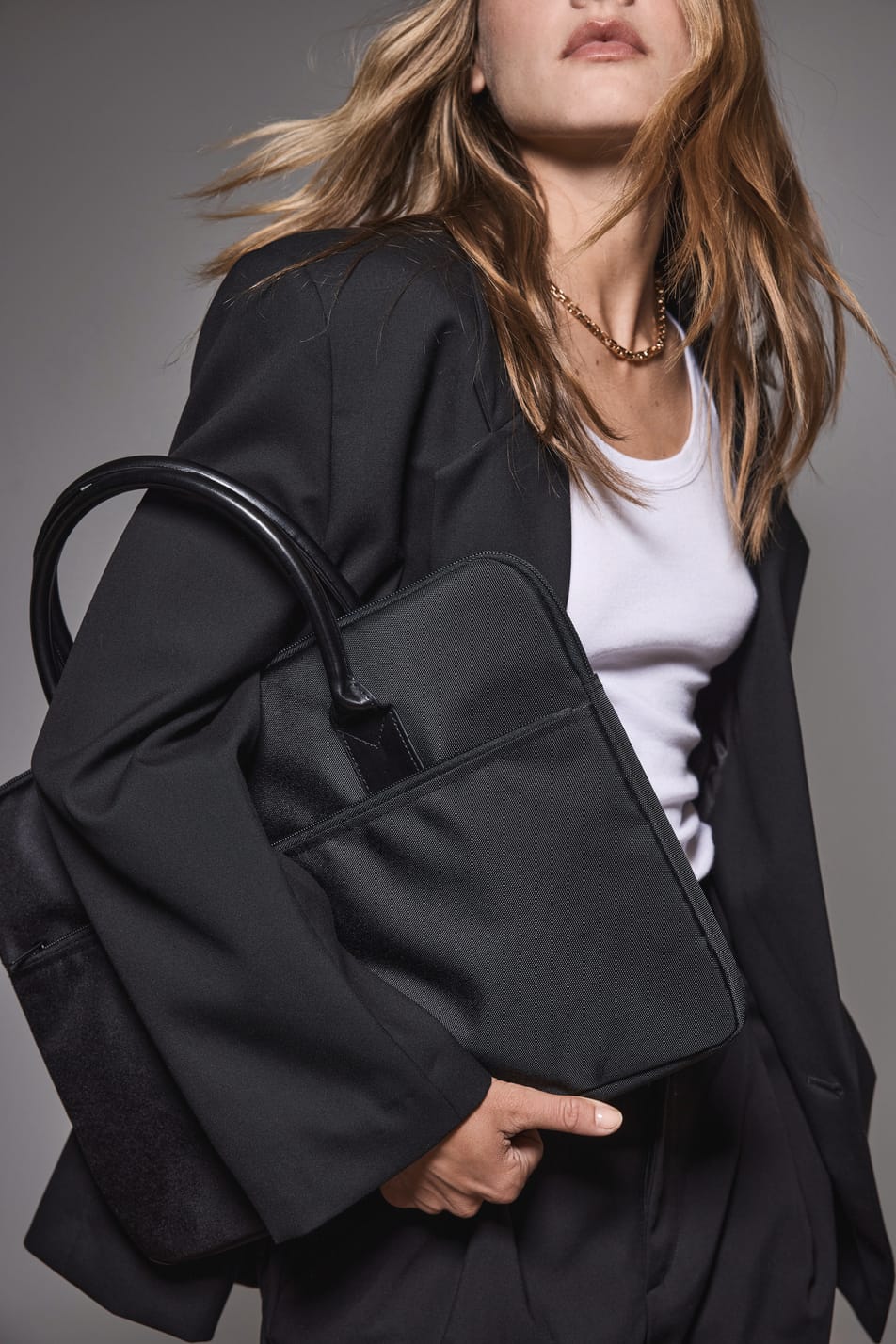 Jeune femme blonde aux cheveux longs en tenue business noire et blanche et tenant un sac porte-ordianteur noir.