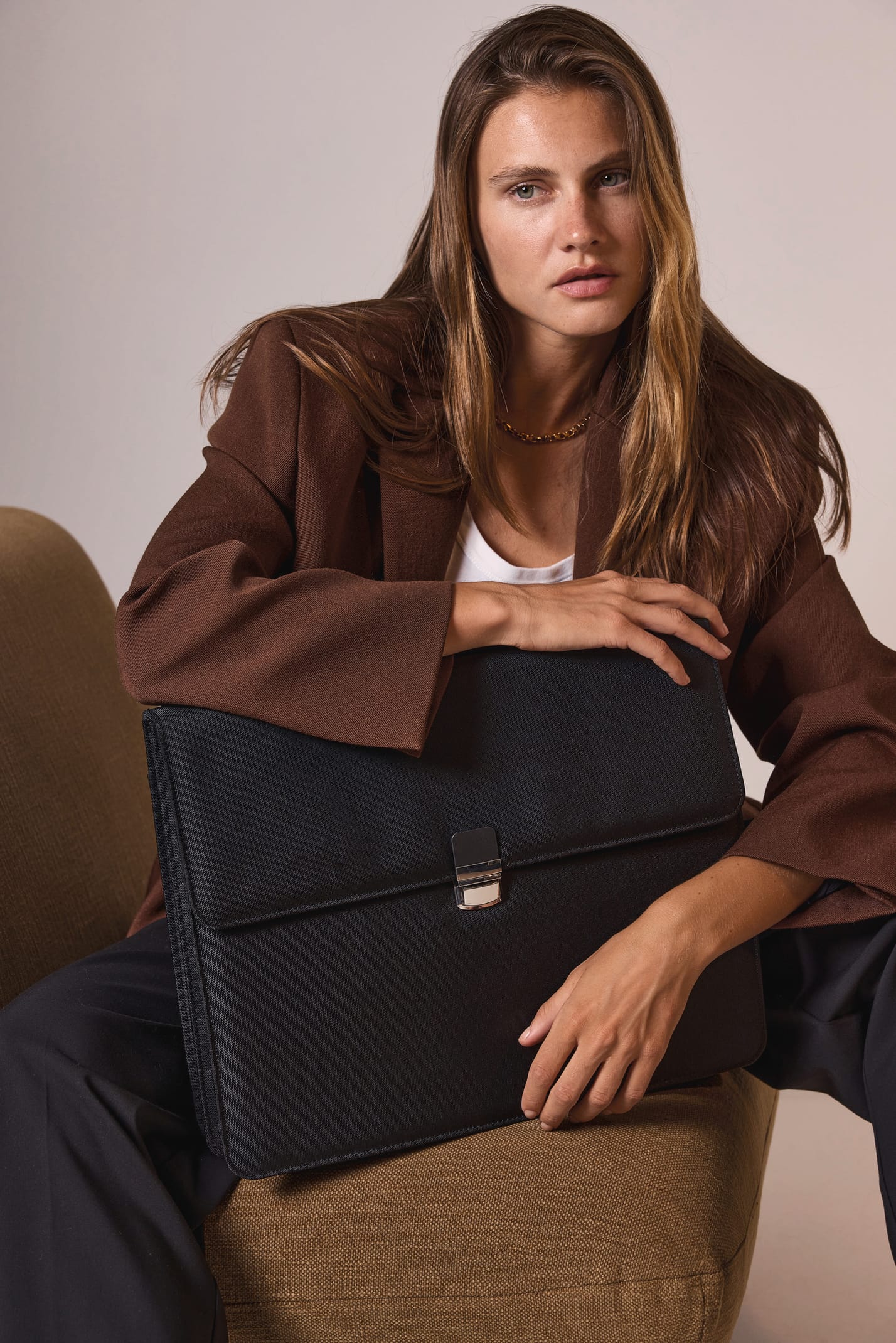 Femme d'affaire blonde aux cheveux long avec une veste chocolat assise  sur un fauteuil marron clair avec un sac noir porte-documents.