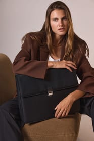 Femme d'affaire blonde aux cheveux long avec une veste chocolat assise  sur un fauteuil marron clair avec un sac noir porte-documents.