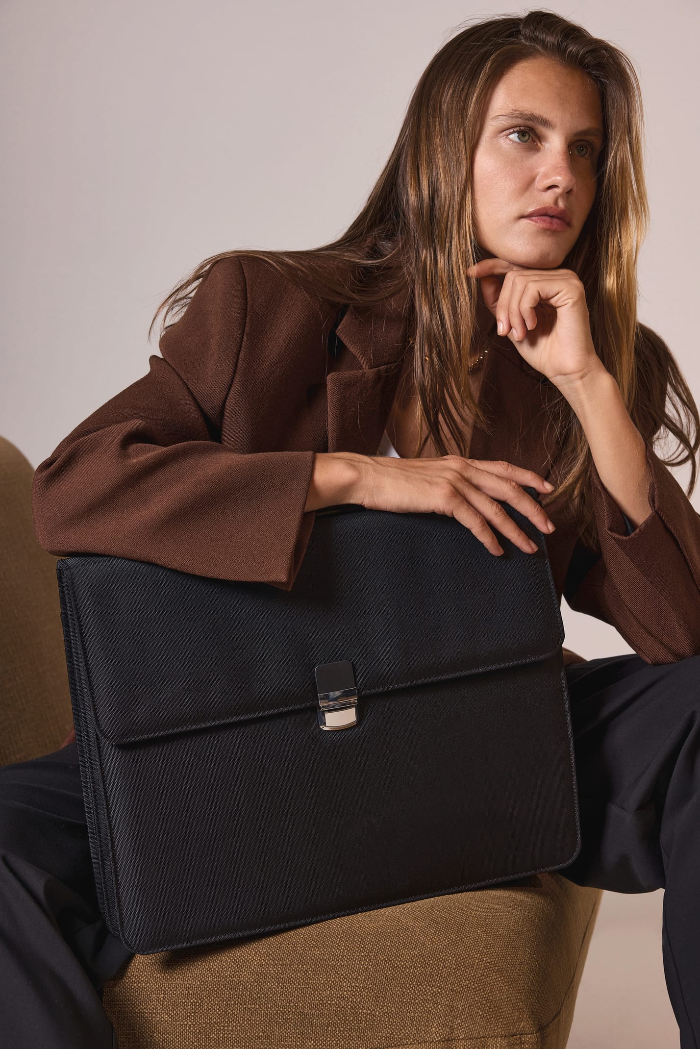 Femme d'affaire blonde aux cheveux long avec une veste chocolat assise  sur un fauteuil marron clair avec un sac noir porte-documents.