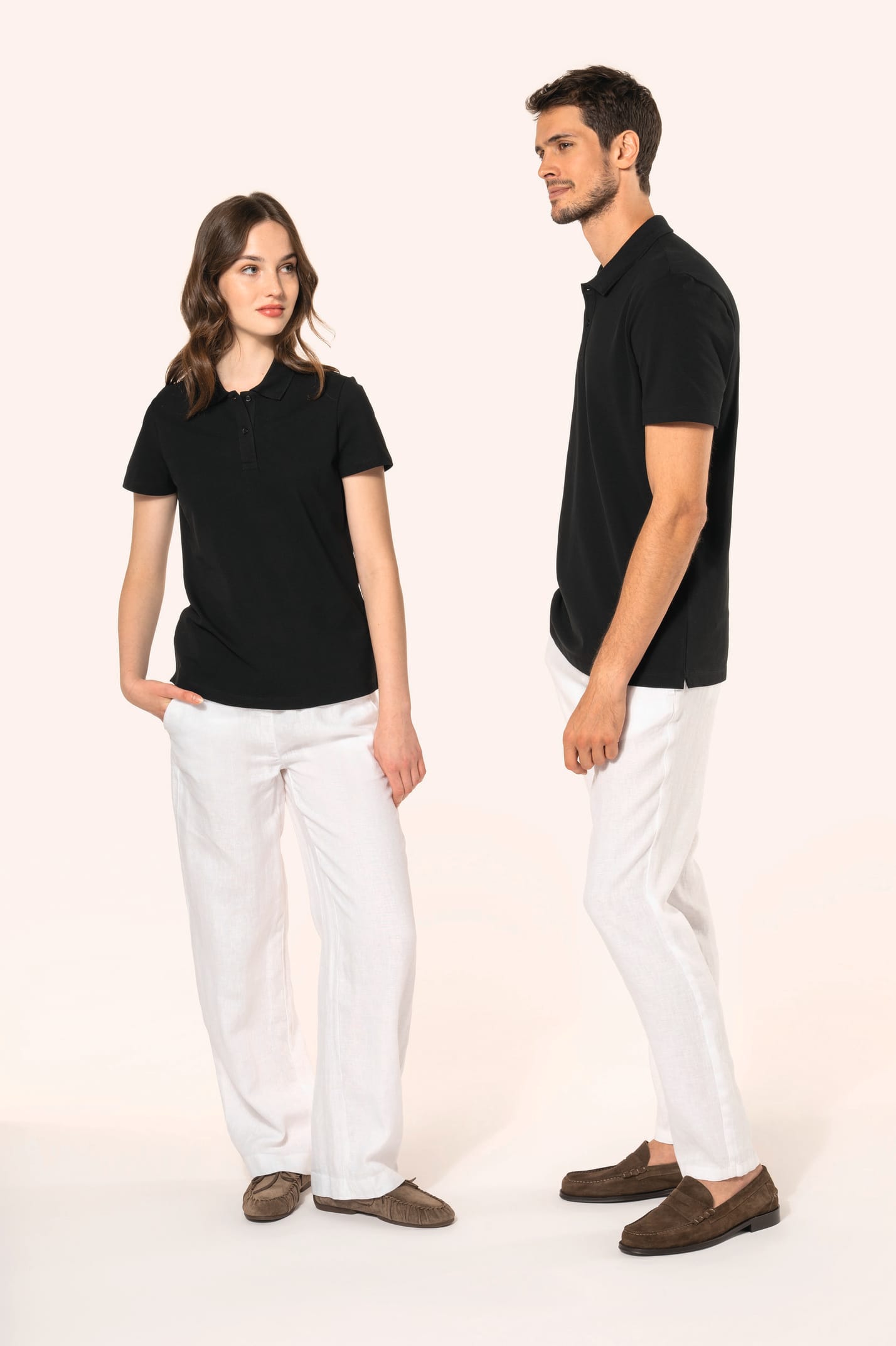 Jeune femme aux cheveux châtains longs et légèrement ondulés, et jeune homme aux cheveux châtains courts portent chacun un polo noir en coton Supima® à manches courtes, sobre et élégant. Sa fibre extra-longue apporte résistance et douceur, idéale pour le 