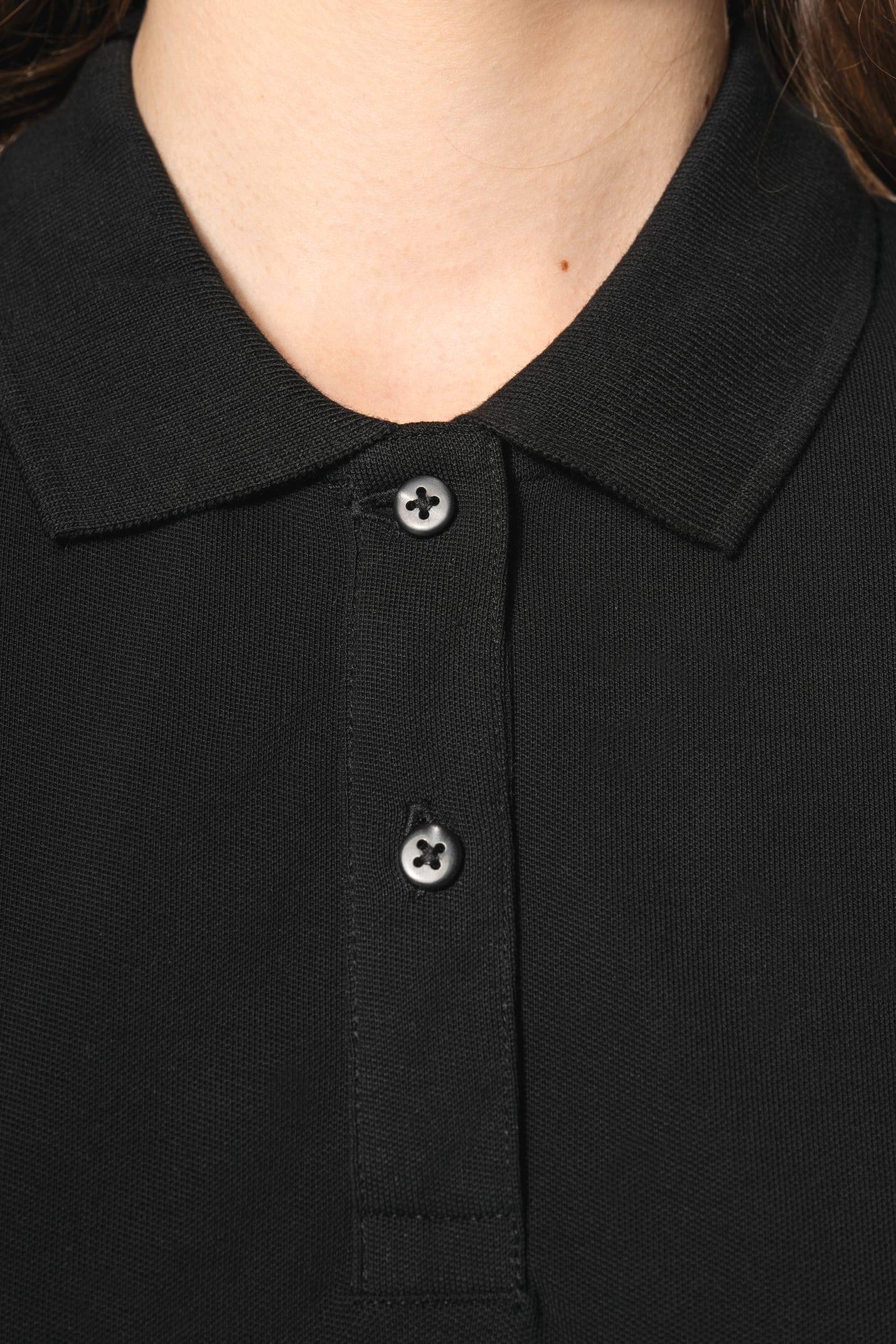Jeune femme aux cheveux châtains longs et légèrement ondulés, portant un polo noir en coton Supima® à manches courtes, sobre et élégant. Sa fibre extra-longue offre résistance et douceur, idéale pour le marquage textile. Elle complète sa tenue avec un pan