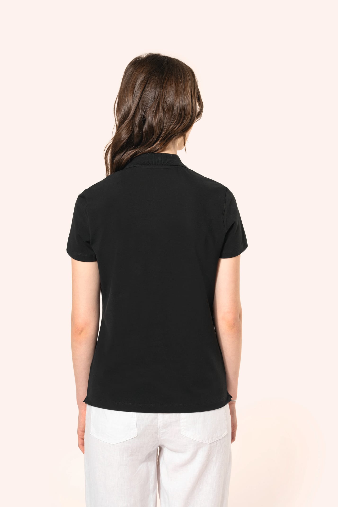 Jeune femme aux cheveux châtains longs et légèrement ondulés, portant un polo noir en coton Supima® à manches courtes, sobre et élégant. Sa fibre extra-longue offre résistance et douceur, idéale pour le marquage textile. Elle complète sa tenue avec un pan