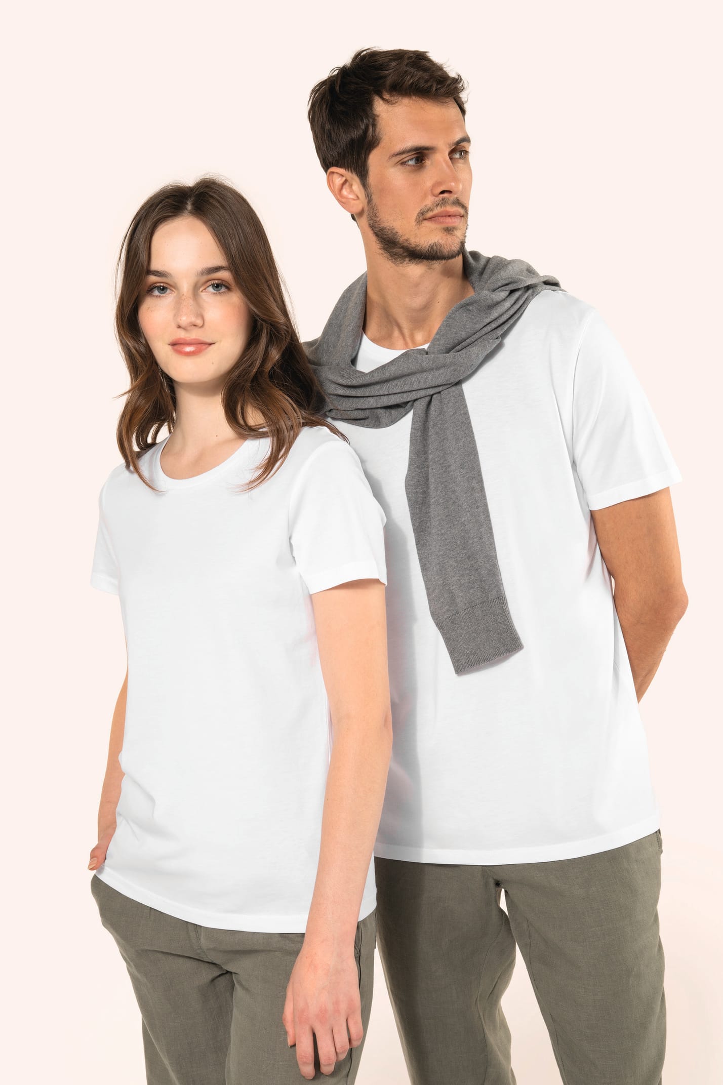 Jeune femme aux cheveux châtains longs et légèrement ondulés et jeune homme aux cheveux châtains courts portent un T-shirt blanc en coton Supima® col rond à manches courtes, 180 g/m². Associé à un pantalon en lin vert kaki clair, l’ensemble met en valeur 