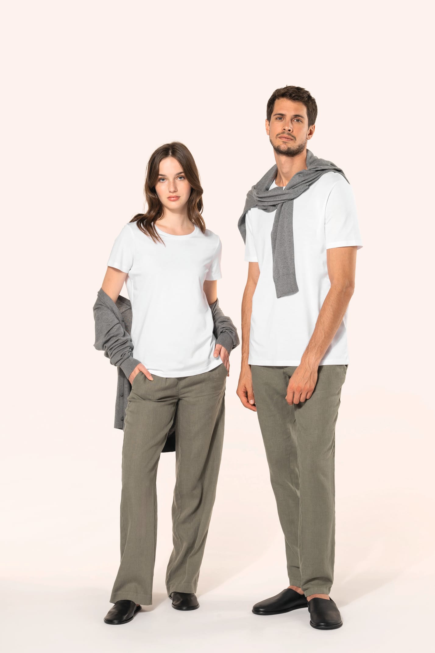 Jeune femme aux cheveux châtains longs et légèrement ondulés et jeune homme aux cheveux châtains courts portent un T-shirt blanc en coton Supima® col rond à manches courtes, 180 g/m². Associé à un pantalon en lin vert kaki clair, l’ensemble met en valeur 