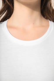 Jeune femme aux cheveux châtains longs et légèrement ondulés portant un T-shirt blanc en coton Supima® col rond à manches courtes, 180 g/m². Associé à un pantalon en lin vert kaki clair et à des chaussures en cuir noir brillantes, l’ensemble met en valeur