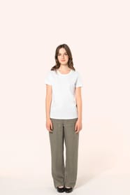 Jeune femme aux cheveux châtains longs et légèrement ondulés portant un T-shirt blanc en coton Supima® col rond à manches courtes, 180 g/m². Associé à un pantalon en lin vert kaki clair et à des chaussures en cuir noir brillantes, l’ensemble met en valeur