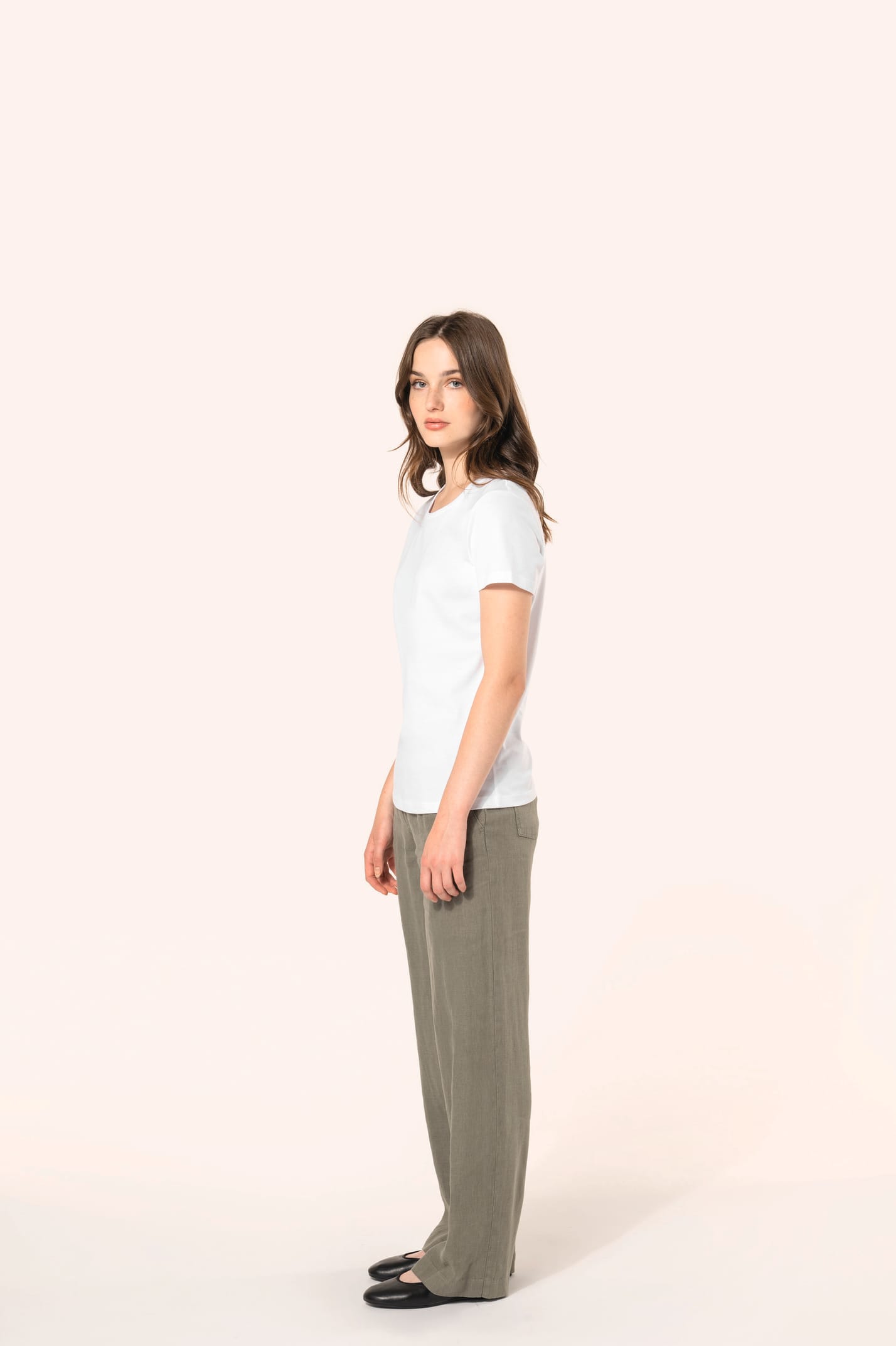 Jeune femme aux cheveux châtains longs et légèrement ondulés portant un T-shirt blanc en coton Supima® col rond à manches courtes, 180 g/m². Associé à un pantalon en lin vert kaki clair et à des chaussures en cuir noir brillantes, l’ensemble met en valeur
