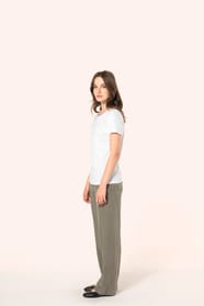 Jeune femme aux cheveux châtains longs et légèrement ondulés portant un T-shirt blanc en coton Supima® col rond à manches courtes, 180 g/m². Associé à un pantalon en lin vert kaki clair et à des chaussures en cuir noir brillantes, l’ensemble met en valeur
