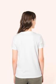 Jeune femme aux cheveux châtains longs et légèrement ondulés portant un T-shirt blanc en coton Supima® col rond à manches courtes, 180 g/m². Associé à un pantalon en lin vert kaki clair et à des chaussures en cuir noir brillantes, l’ensemble met en valeur