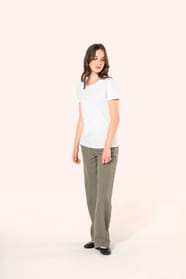 Jeune femme aux cheveux châtains longs et légèrement ondulés portant un T-shirt blanc en coton Supima® col rond à manches courtes, 180 g/m². Associé à un pantalon en lin vert kaki clair et à des chaussures en cuir noir brillantes, l’ensemble met en valeur