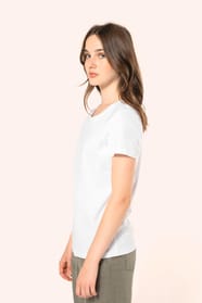 Jeune femme aux cheveux châtains longs et légèrement ondulés portant un T-shirt blanc en coton Supima® col rond à manches courtes, 180 g/m². Associé à un pantalon en lin vert kaki clair et à des chaussures en cuir noir brillantes, l’ensemble met en valeur