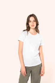 Jeune femme aux cheveux châtains longs et légèrement ondulés portant un T-shirt blanc en coton Supima® col rond à manches courtes, 180 g/m². Associé à un pantalon en lin vert kaki clair et à des chaussures en cuir noir brillantes, l’ensemble met en valeur