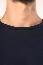 Jeune homme aux cheveux châtains courts portant un T-shirt bleu marine à manches longues en coton Supima® col rond, coupe droite, 180 g/m². Sa fibre extra-longue apporte résistance et douceur, tandis que les finitions soignées et la construction coupée-co