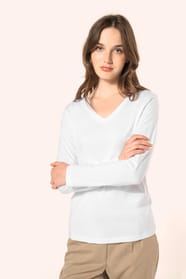 Jeune femme aux cheveux châtains longs et légèrement ondulés portant un T-shirt blanc à manches longues en coton Supima® col V, coupe droite, 180 g/m². Sa fibre extra-longue apporte résistance et douceur, tandis que les finitions soignées et la constructi