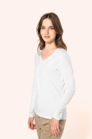Jeune femme aux cheveux châtains longs et légèrement ondulés portant un T-shirt blanc à manches longues en coton Supima® col V, coupe droite, 180 g/m². Sa fibre extra-longue apporte résistance et douceur, tandis que les finitions soignées et la constructi