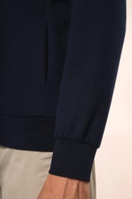 Jeune homme aux cheveux châtains courts portant une veste zippée bleu marine, coupe contemporaine et confortable, 320 g/m². Son tissu bénéficie du traitement LSI (Low Shrinkage Interlock) qui garantit un faible rétrécissement. Il complète sa tenue avec un