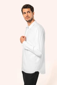 Jeune homme aux cheveux châtains courts portant une chemise blanche en popeline à manches longues, dotée d’un col avec baleines amovibles. Les hirondelles de renfort, petits triangles de tissu reliant les pans avant et arrière, témoignent d’une confection