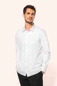 Jeune homme aux cheveux châtains courts portant une chemise blanche en popeline à manches longues, dotée d’un col avec baleines amovibles. Les hirondelles de renfort, petits triangles de tissu reliant les pans avant et arrière, témoignent d’une confection