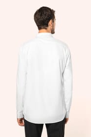 Jeune homme aux cheveux châtains courts portant une chemise blanche en popeline à manches longues, dotée d’un col avec baleines amovibles. Les hirondelles de renfort, petits triangles de tissu reliant les pans avant et arrière, témoignent d’une confection