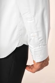 Jeune homme aux cheveux châtains courts portant une chemise blanche en popeline à manches longues, dotée d’un col avec baleines amovibles. Les hirondelles de renfort, petits triangles de tissu reliant les pans avant et arrière, soulignent la précision d’u