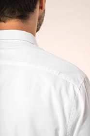 Jeune homme aux cheveux châtains courts portant une chemise blanche en popeline à manches longues, dotée d’un col avec baleines amovibles. Les hirondelles de renfort, petits triangles de tissu reliant les pans avant et arrière, soulignent la précision d’u