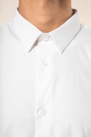 Jeune homme aux cheveux châtains courts portant une chemise blanche en popeline à manches longues, dotée d’un col avec baleines amovibles. Les hirondelles de renfort, petits triangles de tissu reliant les pans avant et arrière, soulignent la précision d’u