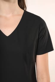 Jeune femme aux cheveux châtains longs attachés en demi-queue portant une robe noire à la coupe cintrée et élégante. Ses finitions soignées apportent classe et sobriété, tandis que son encolure en V avec finition intérieure doublée souligne le raffinement