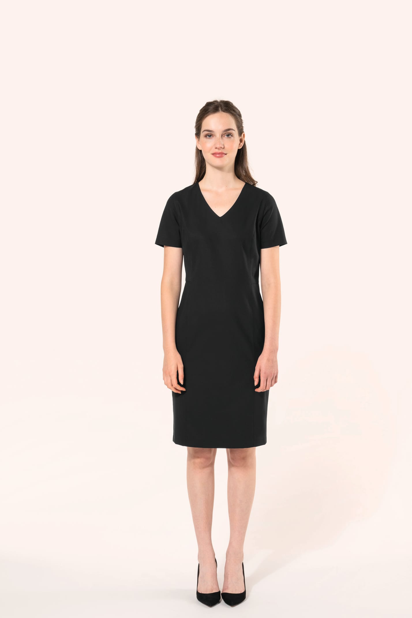 Jeune femme aux cheveux châtains longs attachés en demi-queue portant une robe noire à la coupe cintrée et élégante. Ses finitions soignées apportent classe et sobriété, tandis que son encolure en V avec finition intérieure doublée souligne le raffinement