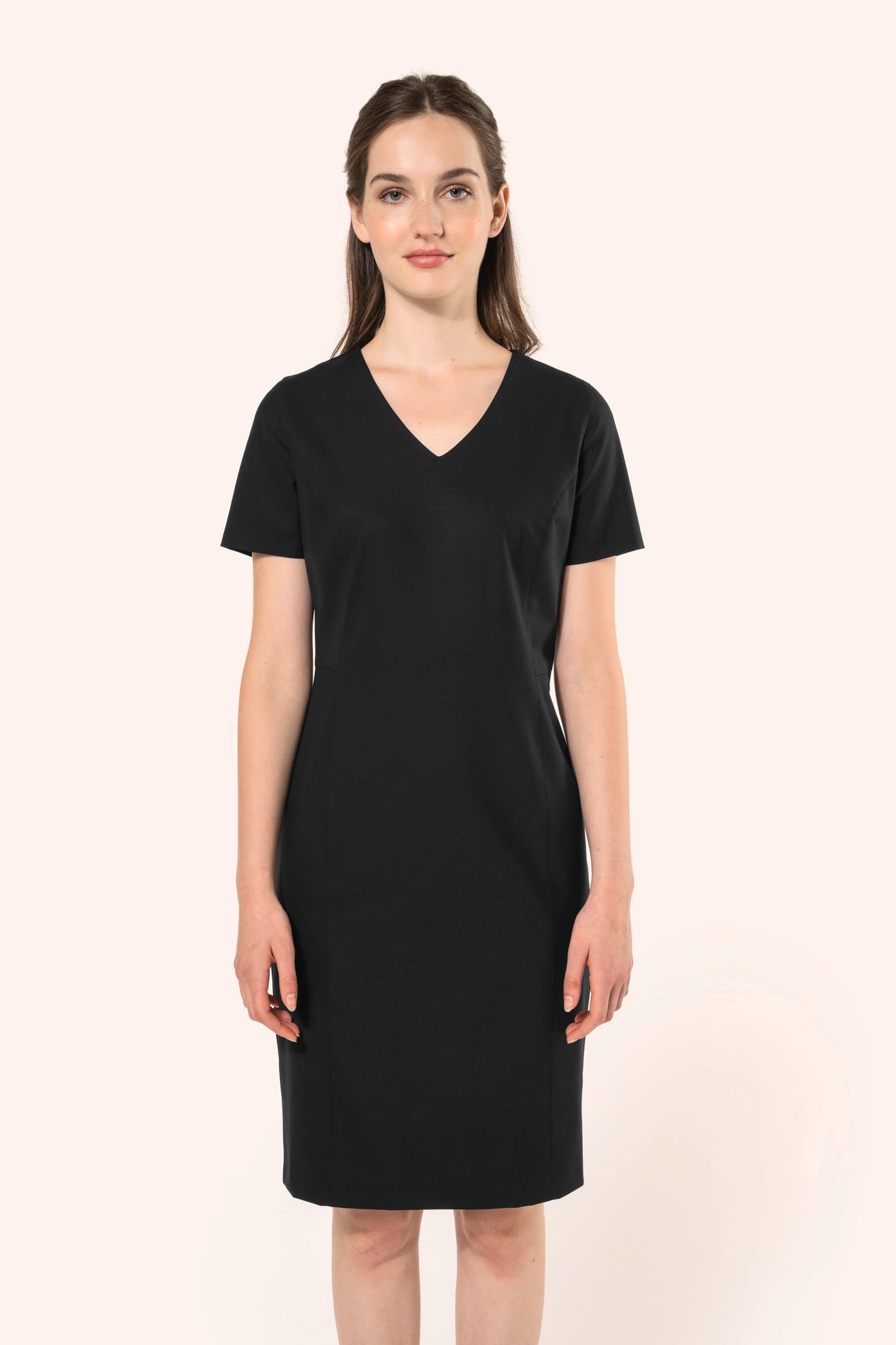 Jeune femme aux cheveux châtains longs attachés en demi-queue portant une robe noire à la coupe cintrée et élégante. Ses finitions soignées apportent classe et sobriété, tandis que son encolure en V avec finition intérieure doublée souligne le raffinement