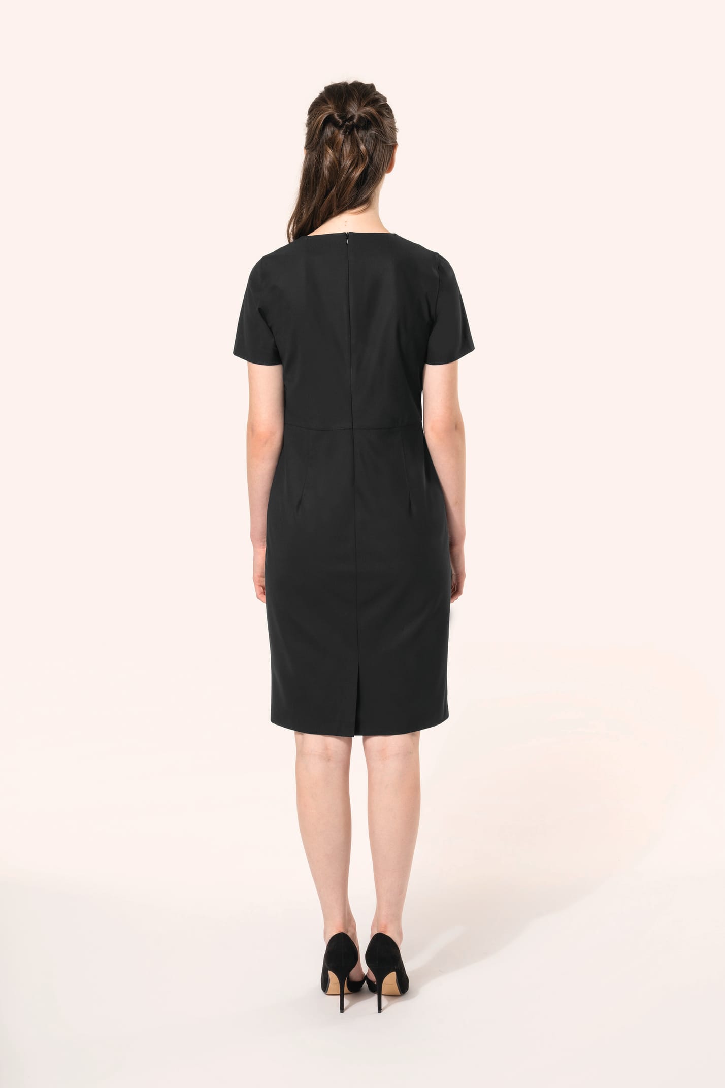 Jeune femme aux cheveux châtains longs attachés en demi-queue portant une robe noire à la coupe cintrée et élégante. Ses finitions soignées apportent classe et sobriété, tandis que son encolure en V avec finition intérieure doublée souligne le raffinement