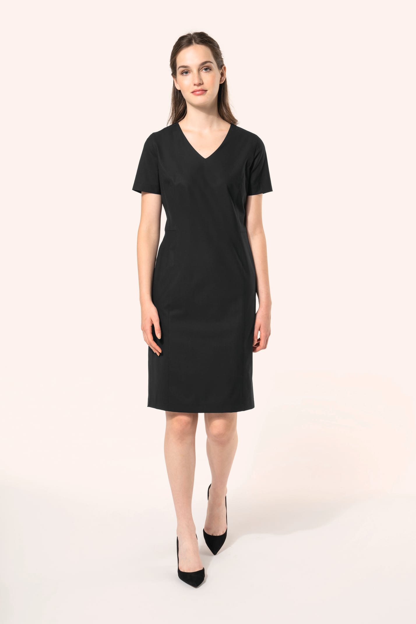 Jeune femme aux cheveux châtains longs attachés en demi-queue portant une robe noire à la coupe cintrée et élégante. Ses finitions soignées apportent classe et sobriété, tandis que son encolure en V avec finition intérieure doublée souligne le raffinement