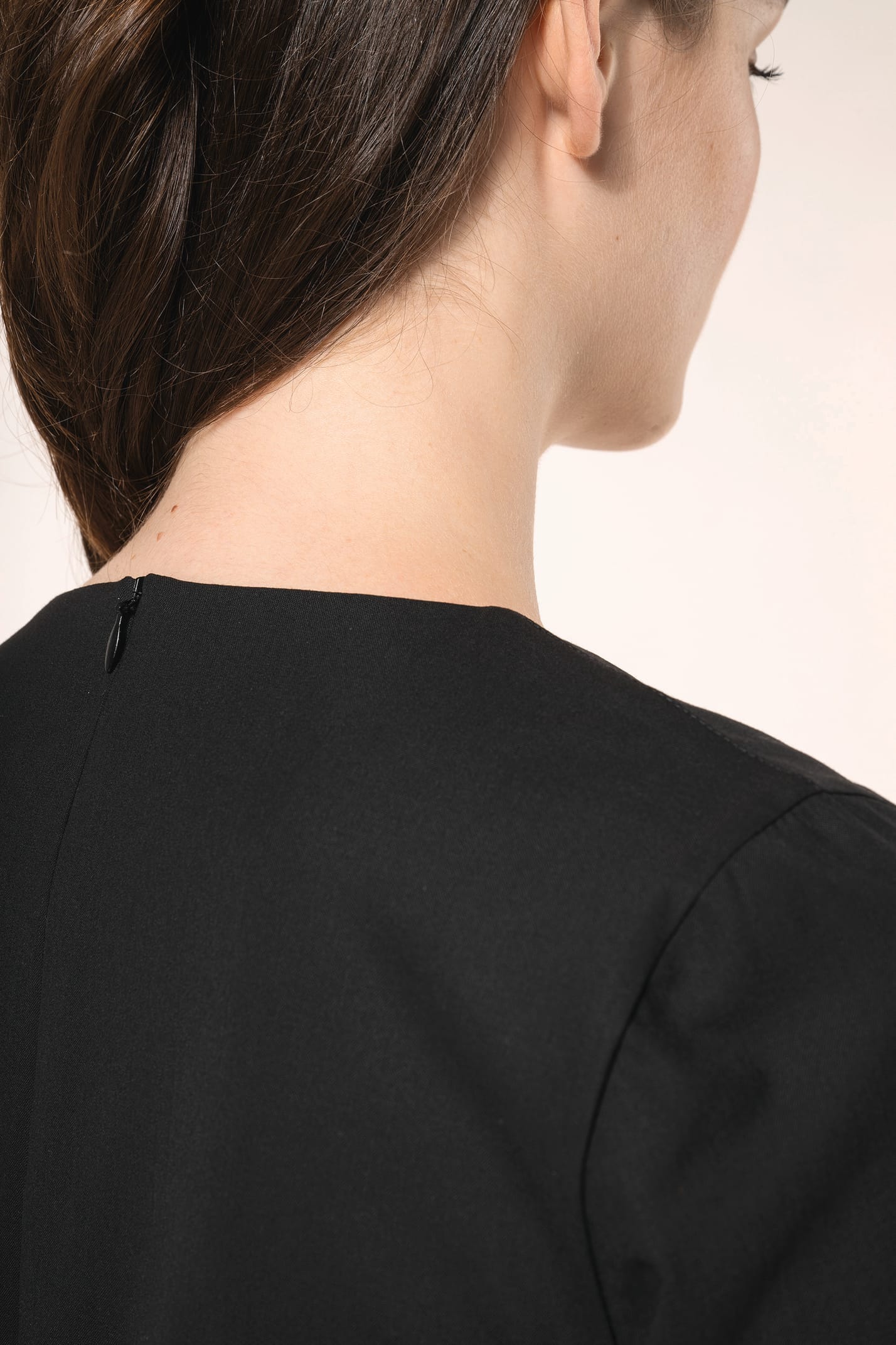 Jeune femme aux cheveux châtains longs attachés en demi-queue portant une robe noire à la coupe cintrée et élégante. Ses finitions soignées apportent classe et sobriété, tandis que son encolure en V avec finition intérieure doublée souligne le raffinement