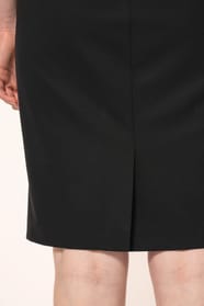 Jeune femme aux cheveux châtains longs attachés en demi-queue portant une robe noire à la coupe cintrée et élégante. Ses finitions soignées apportent classe et sobriété, tandis que son encolure en V avec finition intérieure doublée souligne le raffinement