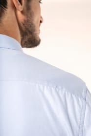 Jeune homme aux cheveux châtains courts portant une chemise bleu clair en popeline à manches longues, confectionnée en coton peigné double retors 80x2 pour une qualité supérieure. Sa coupe ajustée est structurée par deux pinces dans le dos, tandis que les