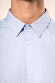 Jeune homme aux cheveux châtains courts portant une chemise bleu clair en popeline à manches longues, confectionnée en coton peigné double retors 80x2 pour une qualité supérieure. Sa coupe ajustée est structurée par deux pinces dans le dos, tandis que les